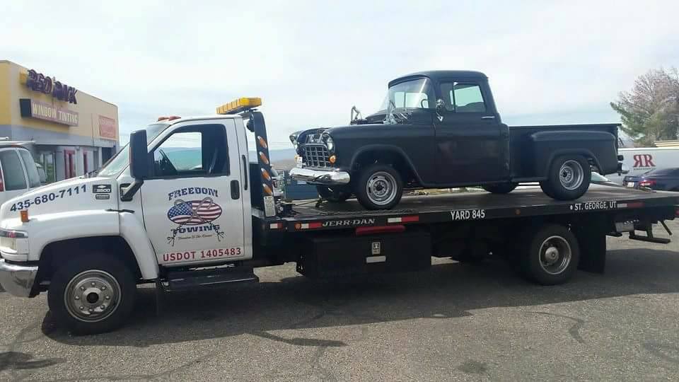 A white Freedom Towing tow truck with a vintage black truck on the bed